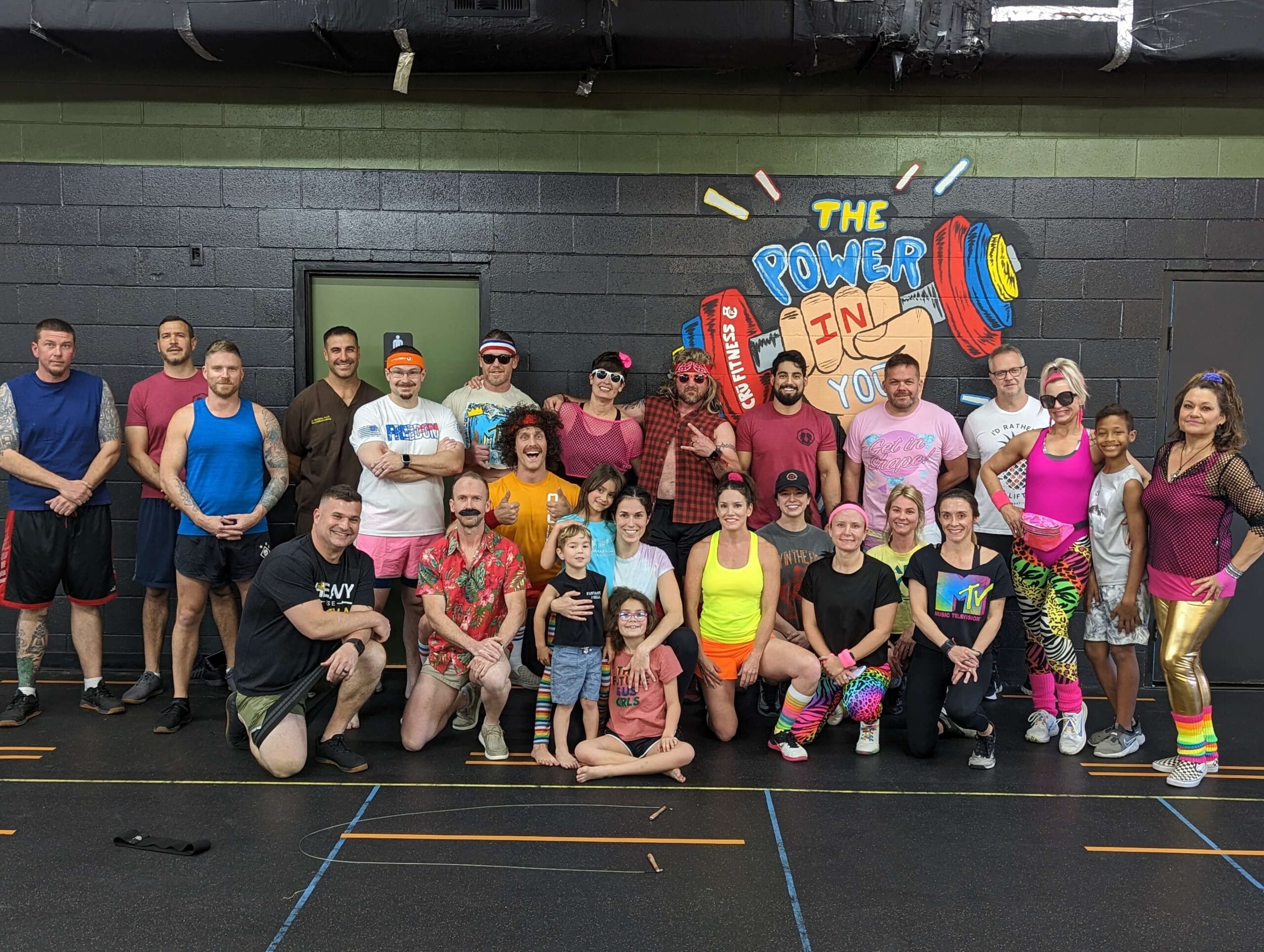 Members and coaches of Cru Fitness pose together in front of mural on the wall at the gym after completing their 80's themed open crossfit workout together